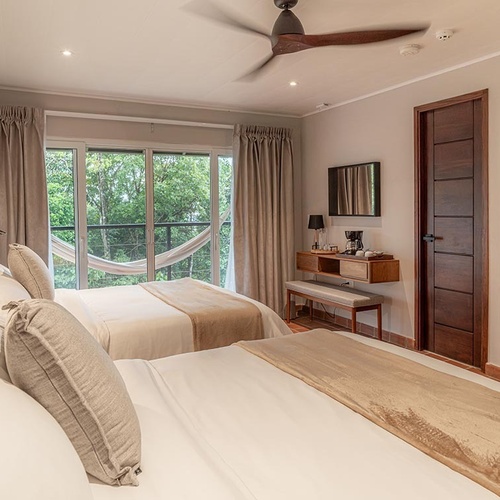 Una luminosa habitación de hotel presenta dos camas, un balcón con hamaca que da a una exuberante vegetación, y un rincón de café junto a una puerta de madera.