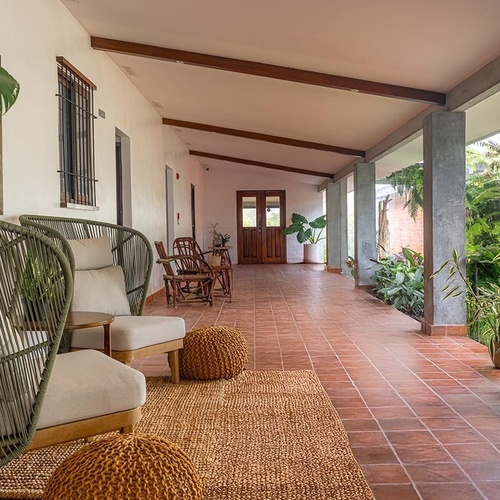 Un amplio corredor exterior con suelo de baldosas de terracota, amueblado con asientos de fibra natural y pufs, que se abre a un exuberante jardín lateral.