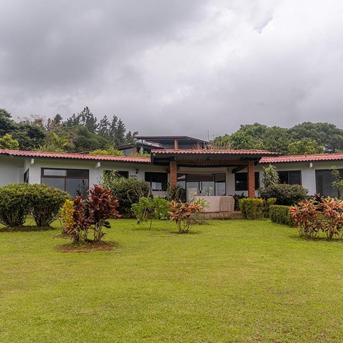 Una casa blanca con techo de tejas rojas y grandes ventanales, rodeada de un césped verde y abundante vegetación, se alza bajo un cielo nublado con un fondo de árboles.