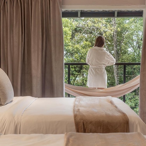 Una persona en bata de baño mira hacia un exuberante bosque desde un balcón con una hamaca, mientras una cama hecha ocupa el primer plano de la habitación.