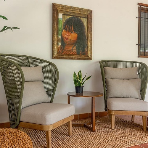 Un acogedor rincón de estar presenta dos sillones de cuerda verde con cojines claros, una mesa auxiliar con una planta, un cuadro de una niña en la pared, una ventana enrejada y detalles rústicos como pufs y una alfombra de yute.