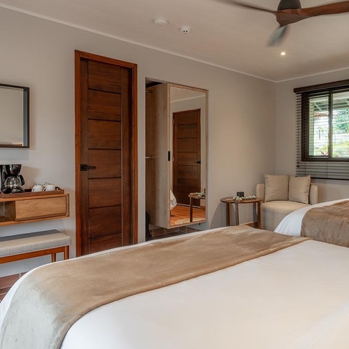 Una acogedora habitación de hotel presenta dos camas con ropa de cama neutra, suelo de baldosas de terracota, muebles de madera oscura y una ventana con persianas.