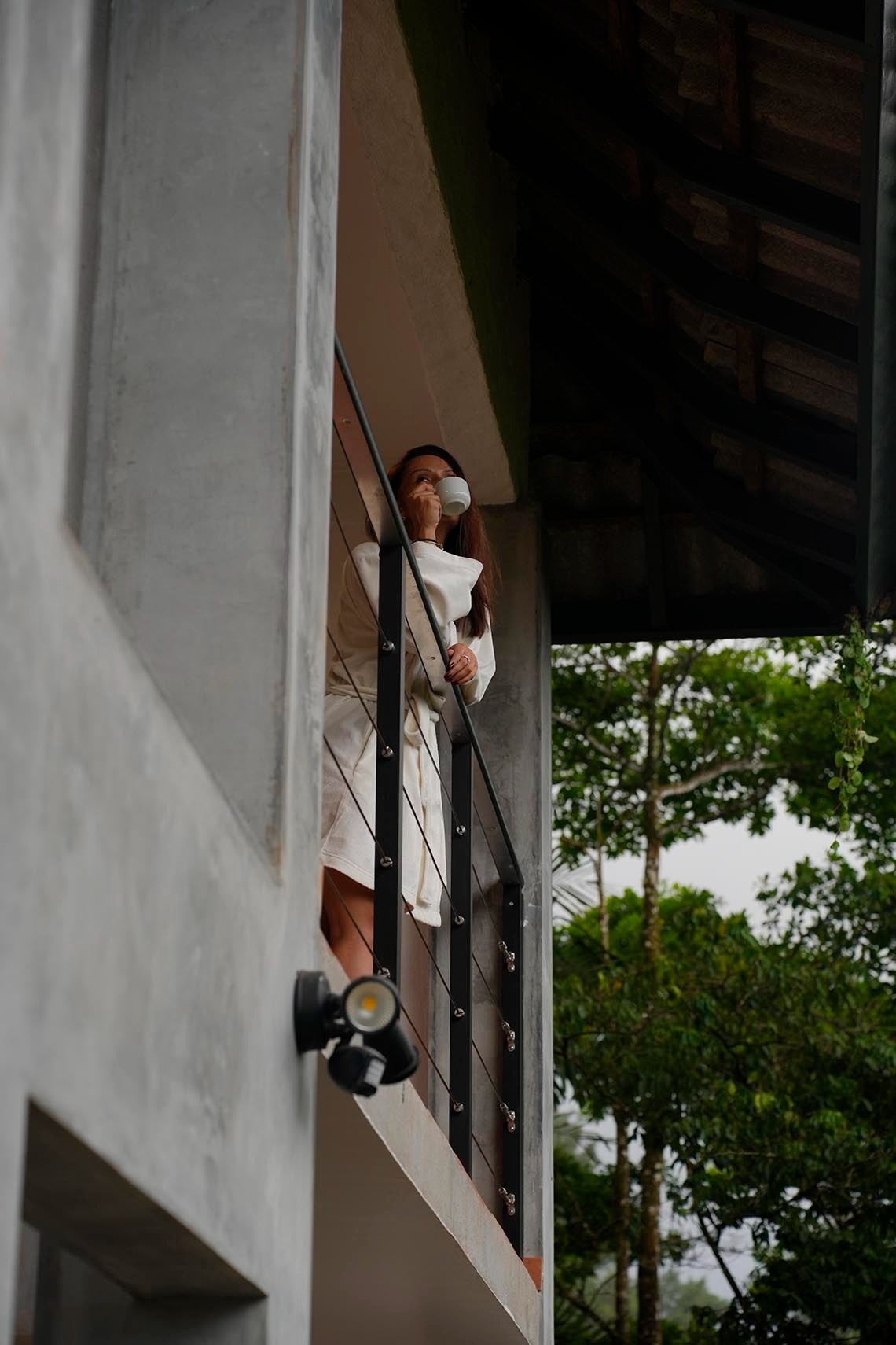 Una mujer en bata beige bebe de una taza mientras mira desde un balcón moderno con barandales de metal y vistas a la vegetación.