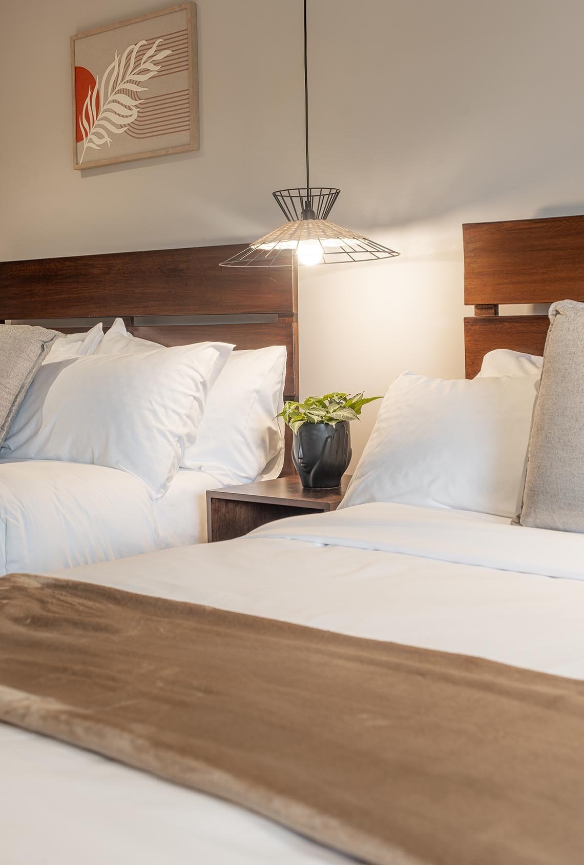 Una habitación moderna y acogedora con dos camas de cabeceros de madera, ropa de cama blanca, una lámpara colgante sobre una mesita de noche con una planta, y un cuadro decorativo en la pared.