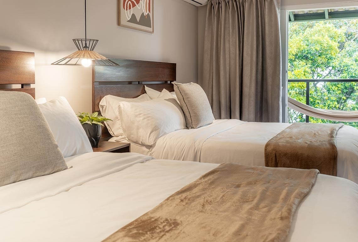 Una habitación de hotel con dos camas de cabeceros de madera, una lámpara colgante de estilo natural y un balcón con hamaca y vistas a la vegetación.