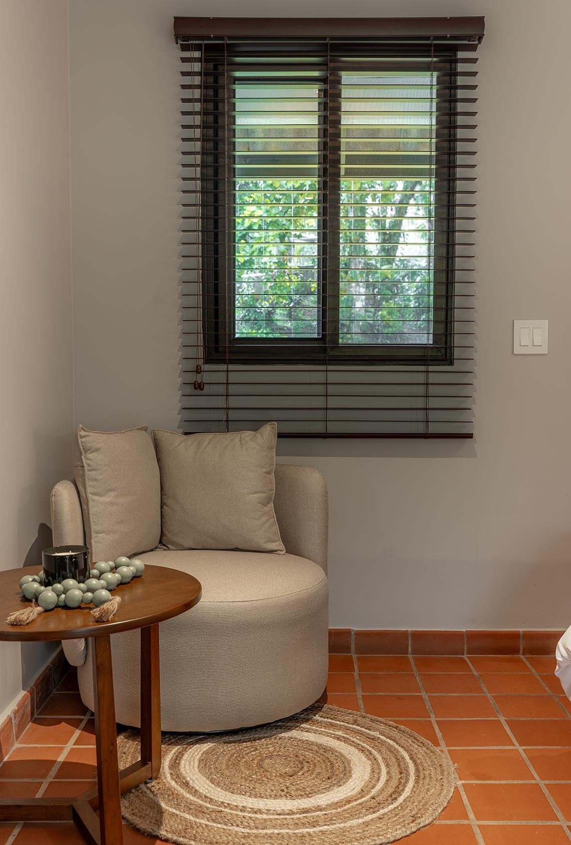 Un rincón acogedor de una habitación presenta una ventana con persianas de madera y vistas a la vegetación exterior, un sillón con cojines, una mesa auxiliar y una alfombra redonda de yute sobre un suelo de baldosas de terracota.