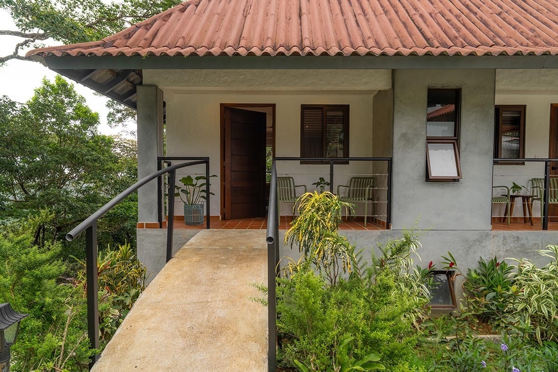 Entrada a una casa con tejado de tejas rojas, una rampa de acceso y un balcón, todo ello rodeado de exuberante vegetación.