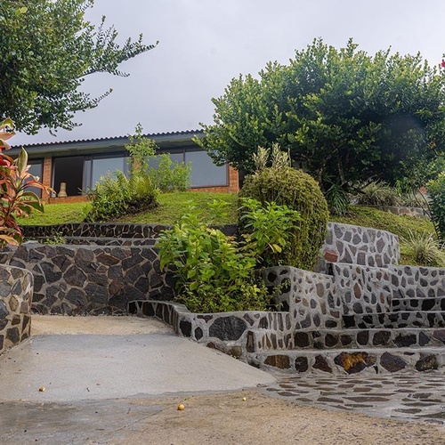Una casa con ventanales se asienta en una colina verde, con escaleras de piedra y exuberante vegetación en primer plano.