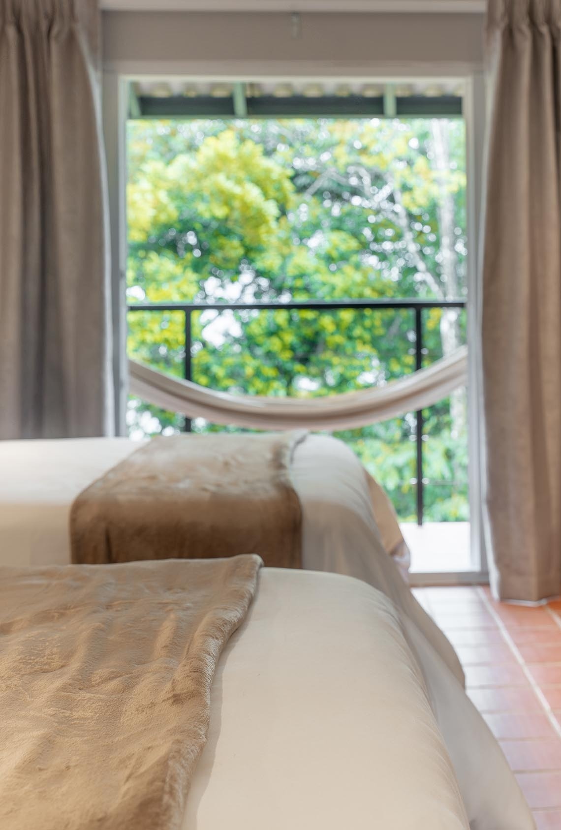 Se ven dos camas con mantas beige frente a un gran ventanal con vistas a la naturaleza verde y un balcón con una hamaca.