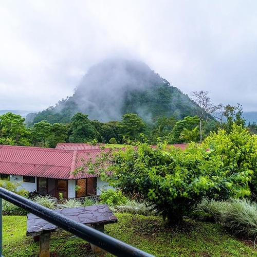 Una vista de un paisaje montañoso con densa vegetación y niebla, donde se aprecian edificios de techos rojos en medio de la naturaleza.
