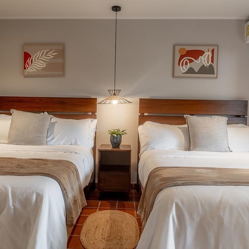 Un dormitorio moderno con dos camas, un velador central con una lámpara colgante, cuadros abstractos en las paredes y suelo de terracota.