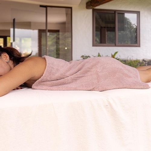 Una mujer yace boca abajo, envuelta en una toalla rosa, sobre una camilla al aire libre.