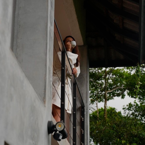 Una mujer con bata blanca bebe de una taza mientras mira pensativamente desde un balcón de hormigón con una barandilla negra, rodeada de árboles verdes.