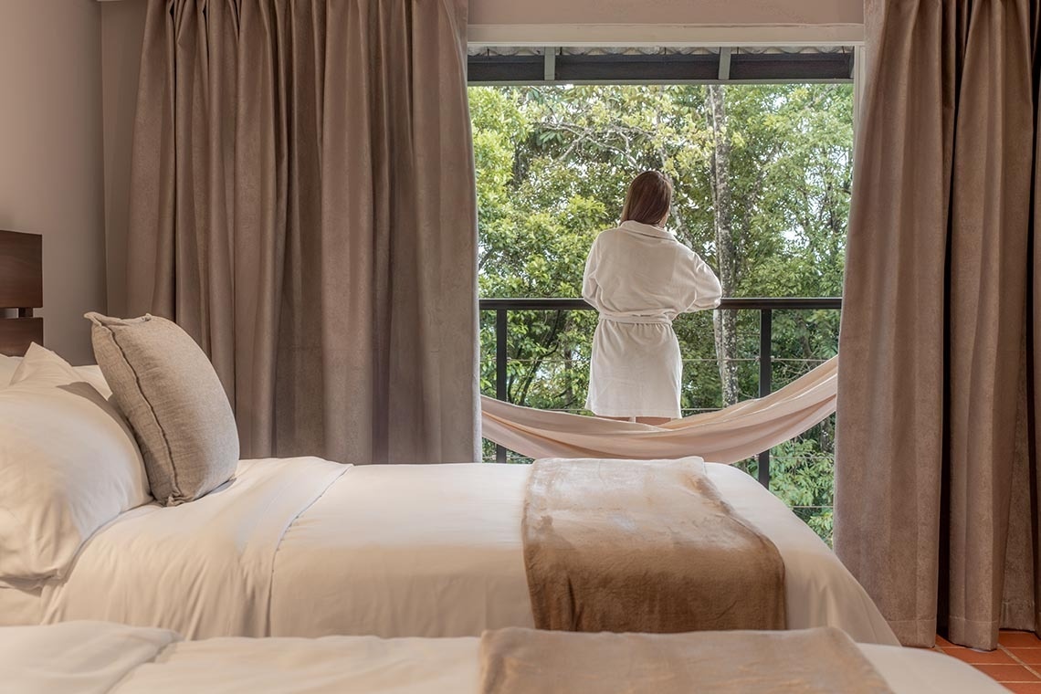 En una habitación con cama y cortinas, una persona en bata blanca observa un bosque frondoso desde un balcón con una hamaca.