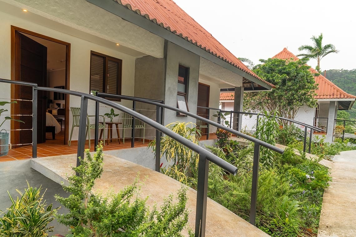 La imagen muestra un complejo de edificios estilo bungalow con techos de teja roja, conectados por rampas y barandillas, en medio de una exuberante vegetación tropical.