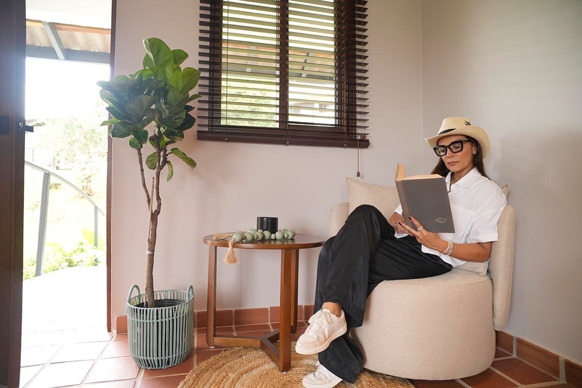 Una mujer con sombrero y gafas está sentada en un sillón leyendo un libro, con una planta y una ventana con persianas de madera de fondo.