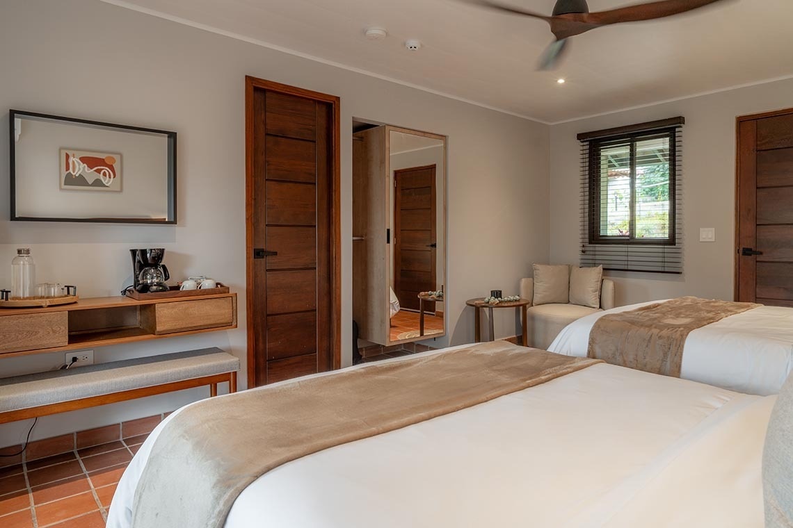 Un dormitorio luminoso de estilo moderno con dos camas, mobiliario y puertas de madera, una estación de café, un ventilador de techo y suelo de baldosas de terracota.