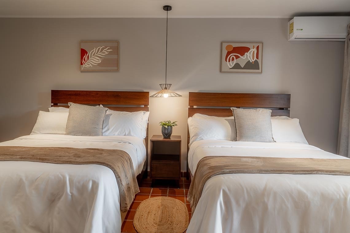 Un dormitorio moderno presenta dos camas con cabeceros de madera, una mesita de noche central con una lámpara colgante y cuadros decorativos en las paredes.