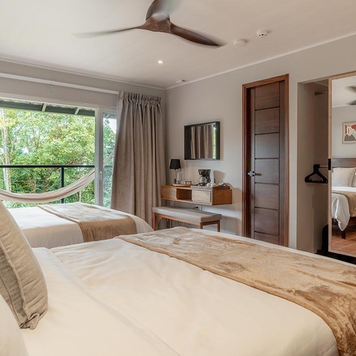 Una habitación luminosa con dos camas, un balcón con hamaca y vistas a la naturaleza, un tocador y un espejo de cuerpo entero.