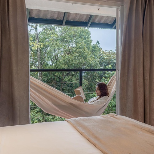 Una mujer lee un libro relajadamente en una hamaca en un balcón con vista a un frondoso bosque, enmarcada por la ventana de una habitación con una cama en primer plano.