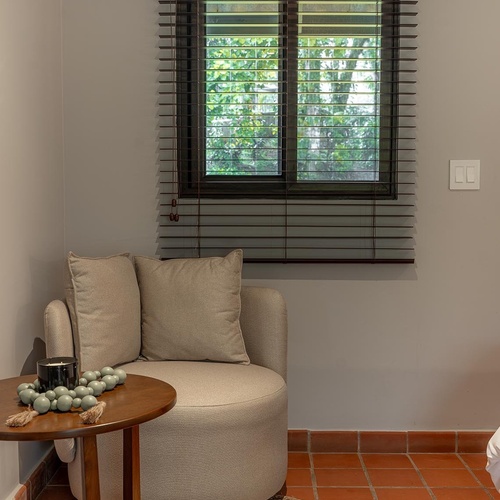Un rincón acogedor cuenta con un sillón beige, una mesa auxiliar de madera con decoración, una alfombra de yute y una ventana con persianas de madera que da a un follaje verde, todo sobre un suelo de baldosas de terracota.