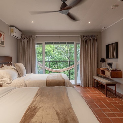 Una habitación de hotel con dos camas, un balcón con vista a la selva y una hamaca, complementada con un ventilador de techo y suelo de baldosas de terracota.
