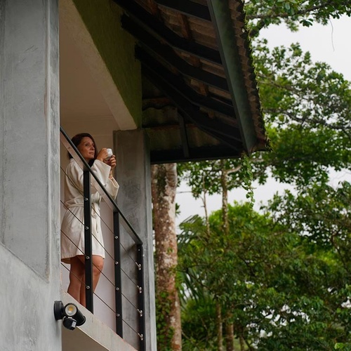 Una mujer en bata de baño está de pie en un balcón, sosteniendo una taza y contemplando el exuberante paisaje verde.
