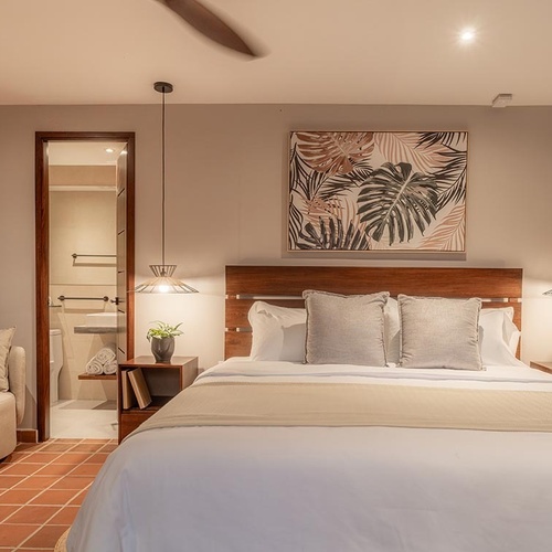 Una habitación moderna y cálida con una cama grande, cabecera de madera, arte tropical en la pared, iluminación ambiental, un área de descanso con sofá y un baño visible, todo sobre un suelo de baldosas de terracota.
