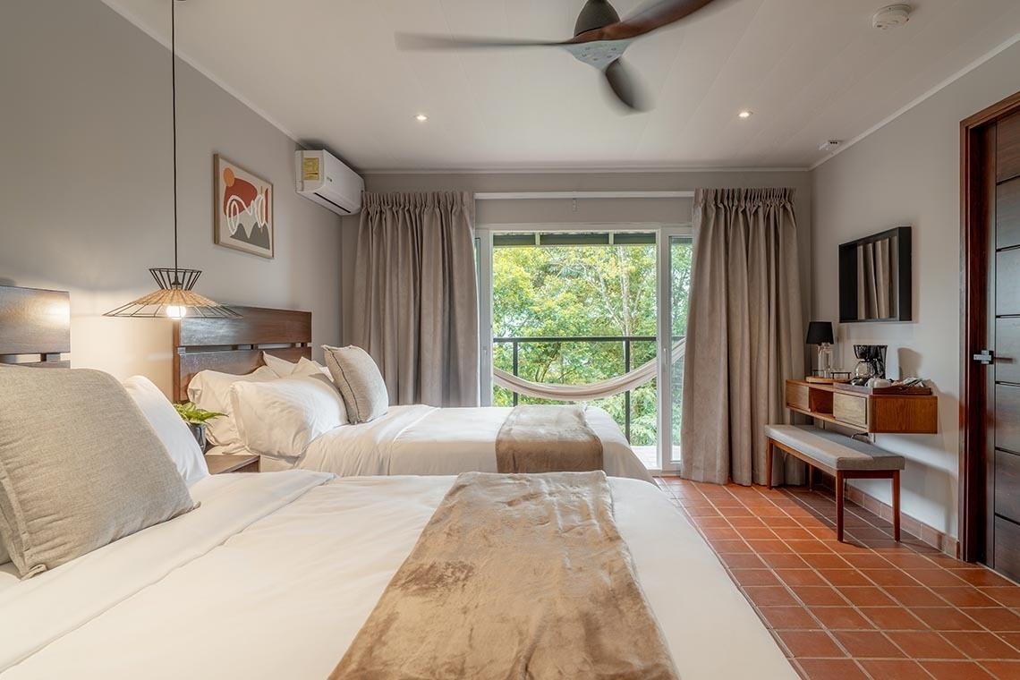 Habitación de hotel con dos camas, balcón con vista al bosque, ventilador de techo y un ambiente acogedor.
