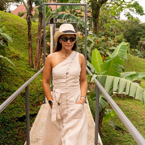 Una mujer con sombrero, gafas de sol y un vestido claro de un hombro camina por una pasarela elevada en un entorno de exuberante vegetación tropical.