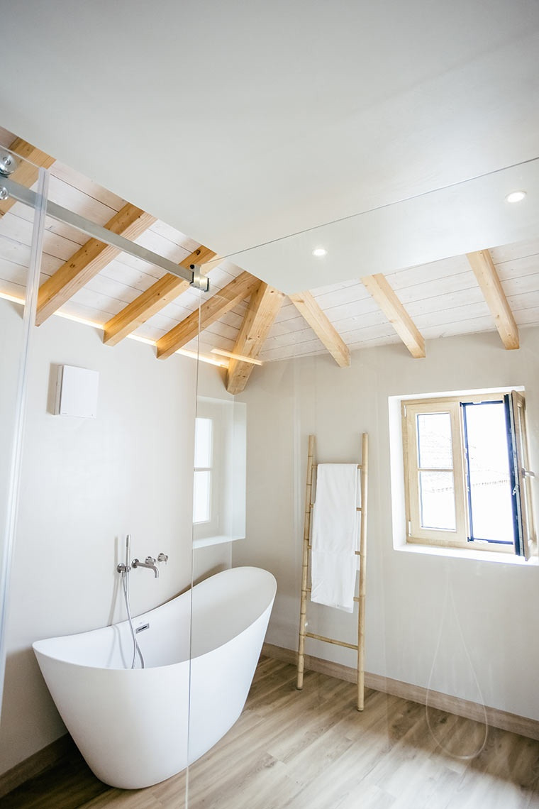 Modern AndTales bathroom with freestanding tub, wood-beamed ceiling, and natural light.