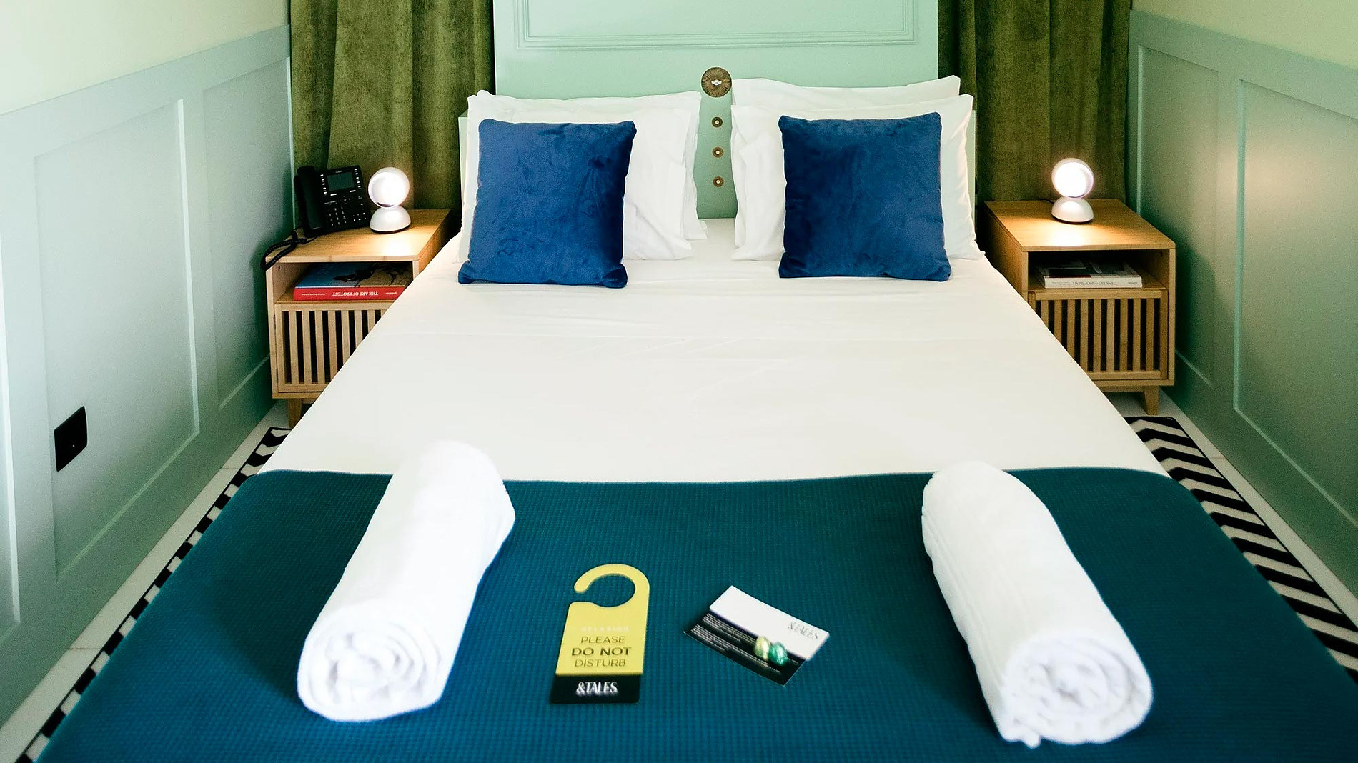 Quarto AndTales: cama com lençóis brancos, almofadas azuis, mantas e porta para não perturbar.