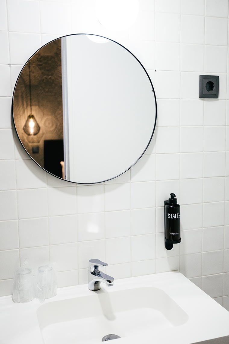 AndTales bathroom sink, mirror, and soap dispenser in a tiled hotel room.