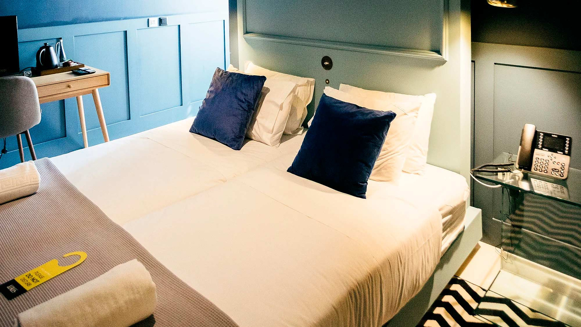 Quarto duplo moderno com duas camas, almofadas azuis e iluminação ambiente no AndTales Hotel.