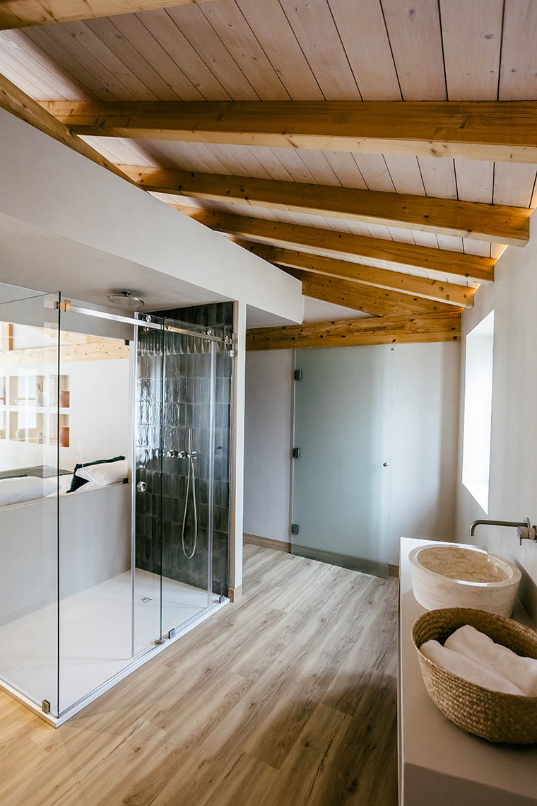 Modern hotel bathroom with glass shower, dual sinks, and rustic AndTales beamed ceiling.
