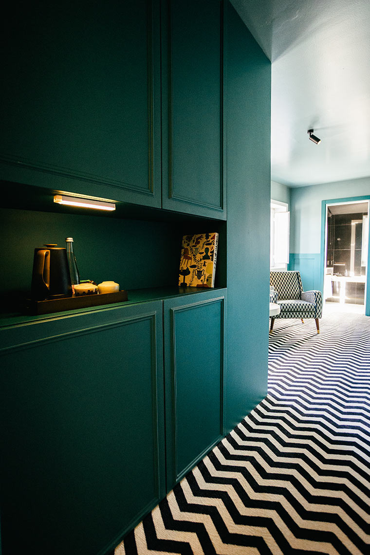 Design do quarto AndTales: móveis verdes escuros, tapete chevron e poltrona.