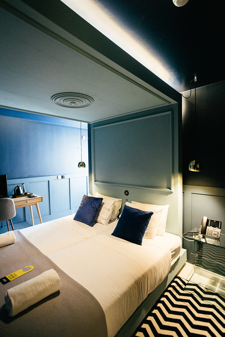 Quarto duplo moderno com duas camas, almofadas azuis e iluminação ambiente no AndTales Hotel.