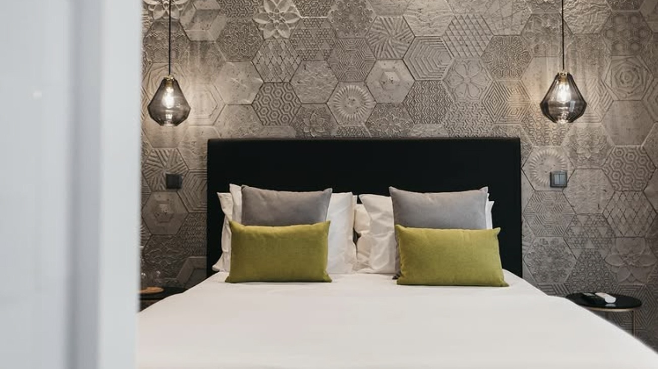 Cama de hotel aconchegante com almofadas verdes e cinzas, e um padrão de azulejo AndTales exclusivo na parede.