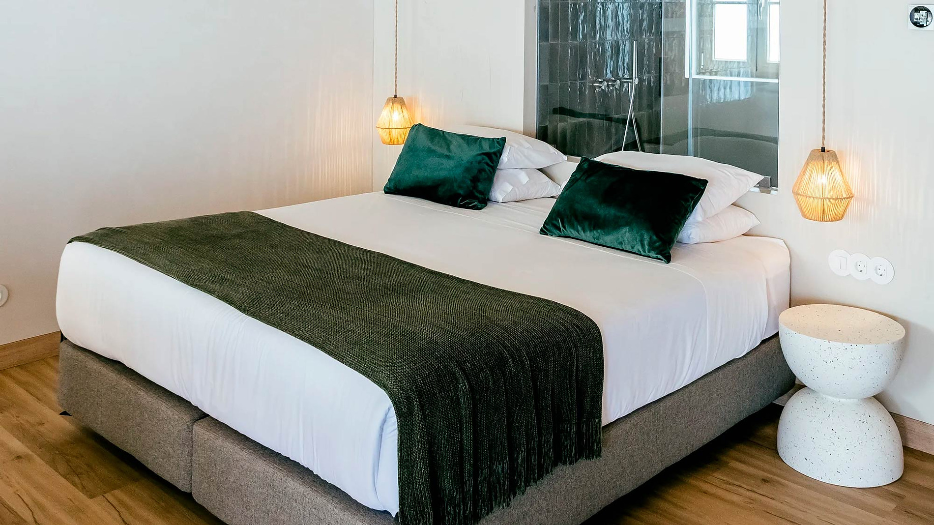 Cama de casal com manta verde e pormenores AndTales num quarto com teto de madeira.
