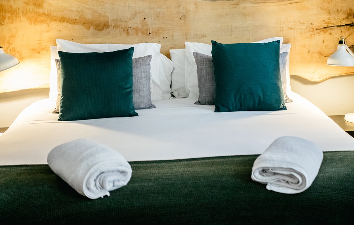 Cama aconchegante em quarto AndTales com travesseiros verdes e mantas enroladas.