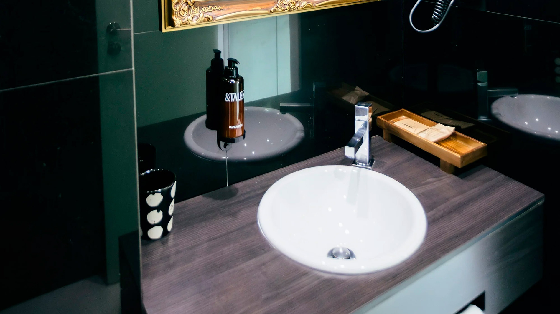 Espelho decorativo em banheiro de hotel AndTales com bancada, pia e secador de cabelo.