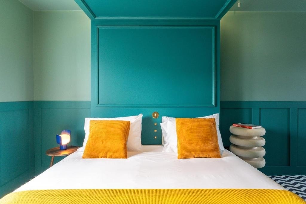 Quarto AndTales: cama com almofadas laranjas, luminária azul e decoração moderna.