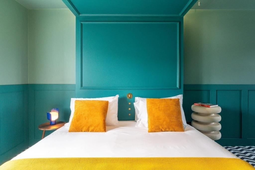 Quarto AndTales: cama com almofadas laranjas, luminária azul e decoração moderna.