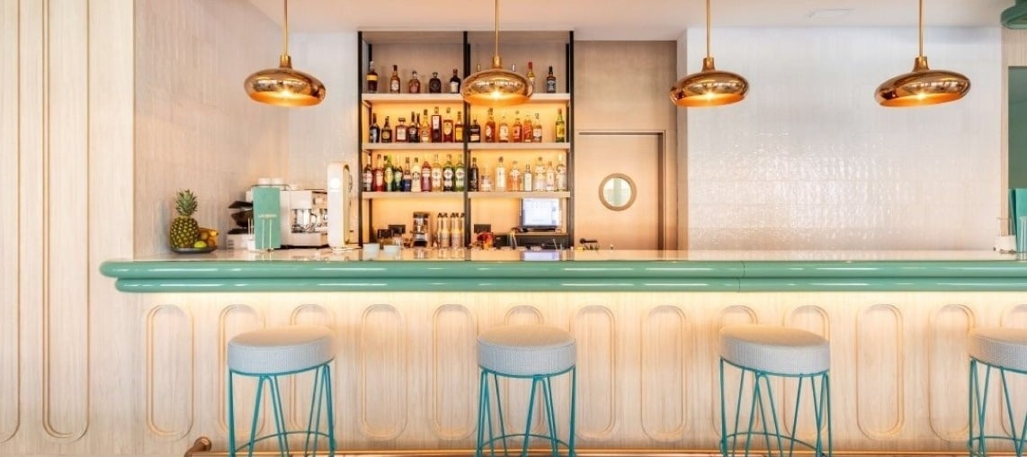 Una imagen de un bar contemporáneo con una barra verde menta iluminada, taburetes a juego, estanterías llenas de botellas de licor y lámparas colgantes doradas.