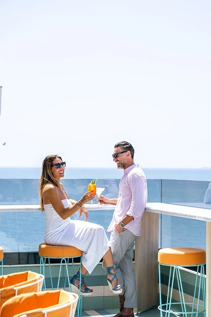 Pareja feliz disfrutando cócteles Angela en terraza con vistas al mar.
