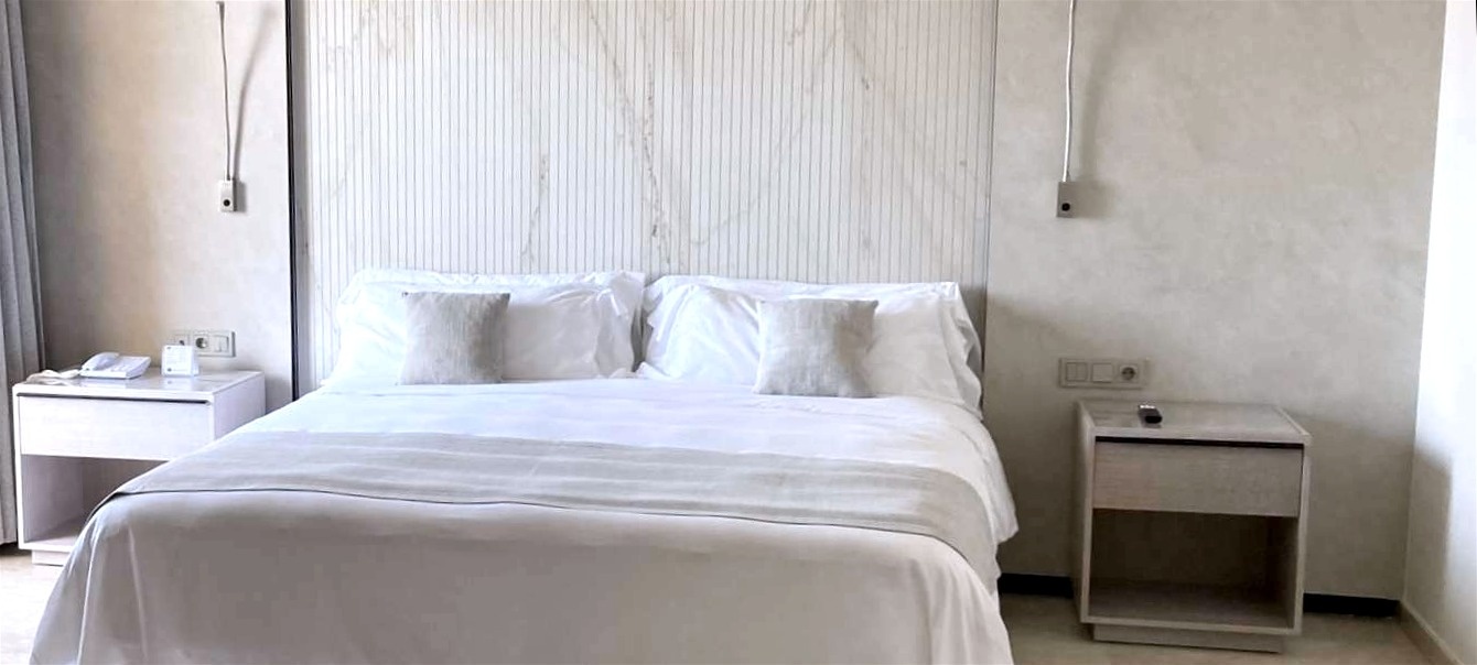 A clean and modern bedroom features a neatly made bed with white linens, flanked by two light-toned nightstands and wall-mounted lamps, set against a textured headboard with vertical grooves and subtle marble-like patterns, and light beige walls.