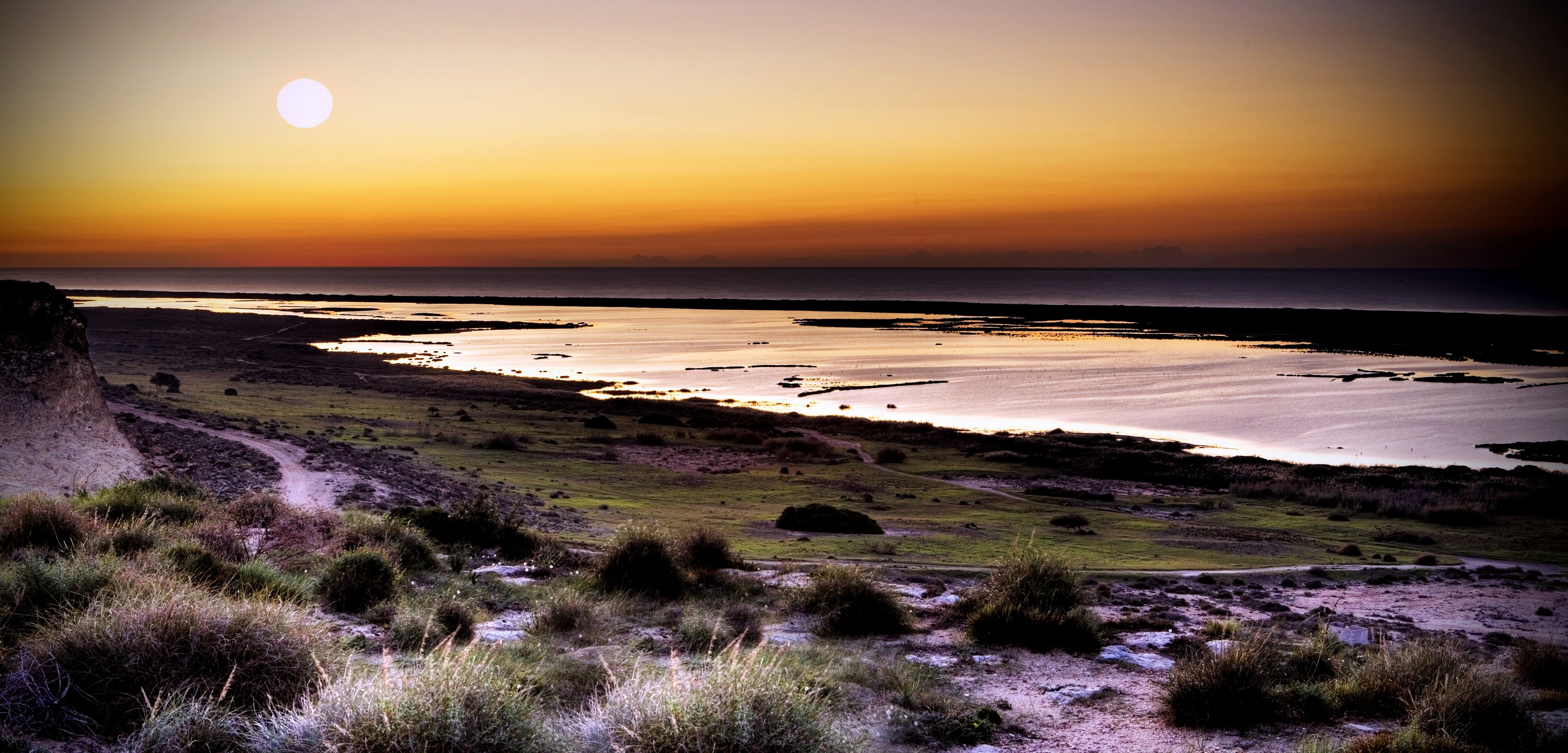 Panorama costero al amanecer o atardecer, con una laguna que refleja el sol bajo sobre una tierra árida y acantilados.