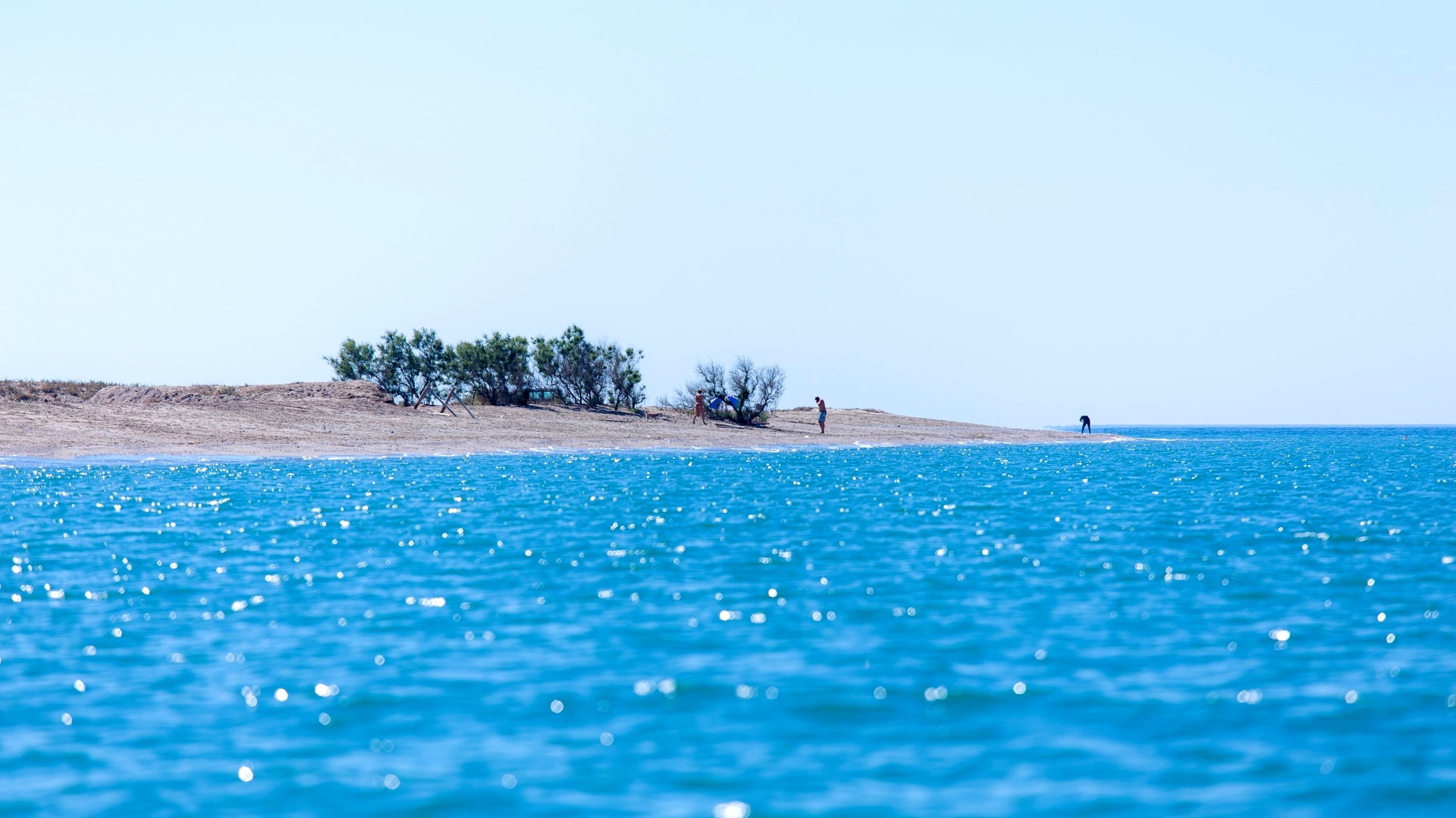 Un paisaje sereno muestra agua azul brillante en primer plano, una orilla arenosa salpicada de vegetación y algunas figuras humanas bajo un cielo despejado.