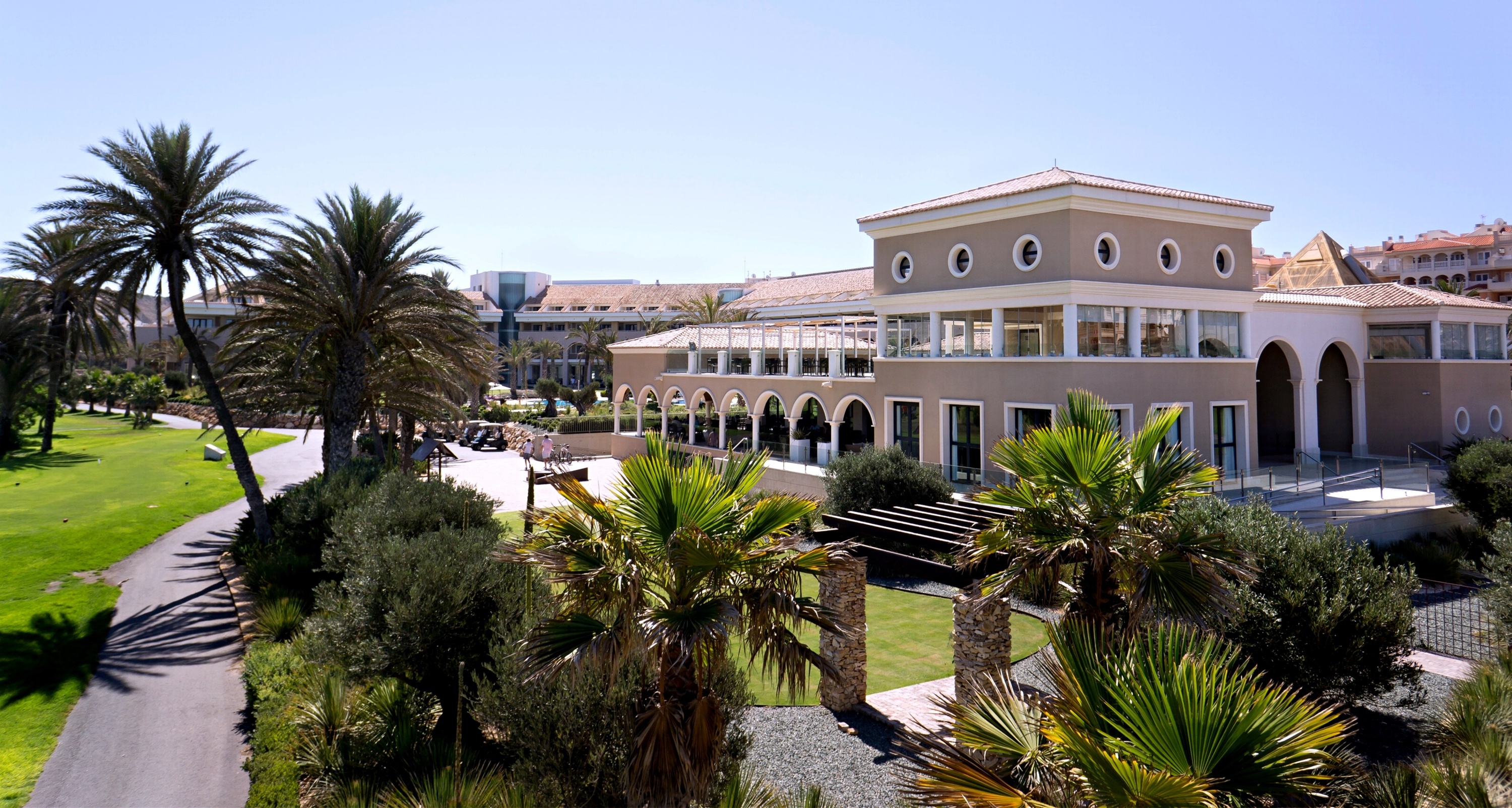 Un edificio de estilo resort con arcos y ventanas redondas se asienta entre palmeras y césped verde, bajo un cielo azul claro y soleado.