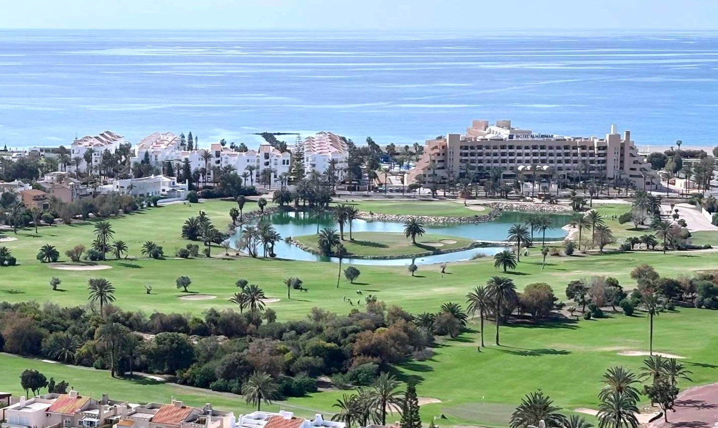 La imagen presenta una vista panorámica de un campo de golf costero con palmeras, un lago, edificios y un hotel frente al mar Mediterráneo.